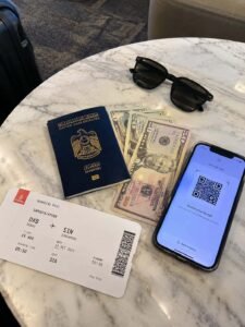 Flat-lay mobile photo of a UAE passport, printed hotel booking voucher, US dollar bills, and boarding pass on a cafe table